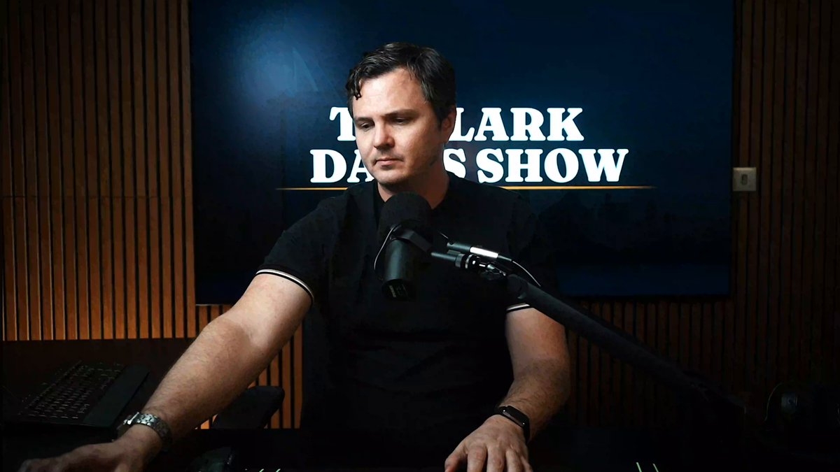 The image features a man sitting at a desk with a microphone, likely preparing for an interview or podcast recording. He appears to be wearing a black shirt as he sits down. In front of him, there's a TV screen displaying "The Clark D. Show". The background is dark and out of focus, drawing attention to the main subject - the man sitting at the desk with his microphone.