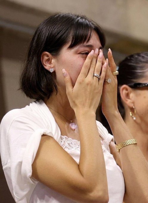 A woman in a white top covers her face with her hands, showing off a diamond ring and a pink heart necklace, appearing emotional.