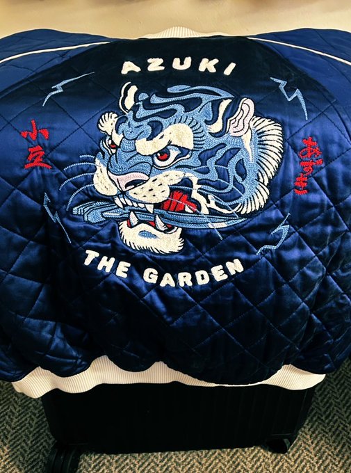 The back of a navy blue satin sukajan-style jacket featuring an embroidered tiger head, with 'AZUKI' embroidered at the top and 'THE GARDEN' at the bottom.