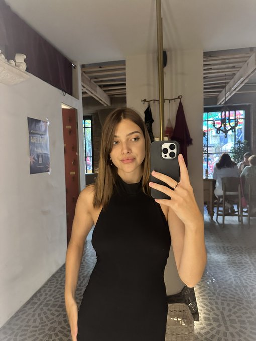 A mirror selfie of a young woman with shoulder-length brown hair wearing a black halter top, holding a smartphone in what appears to be a rustic restaurant or café setting.