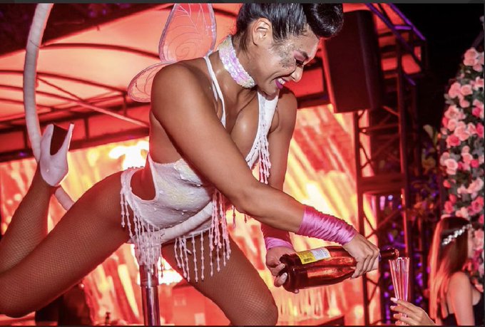 A performer in a sparkly white fairy costume with pink gloves pours from a bottle into a shot glass at a club or event venue.