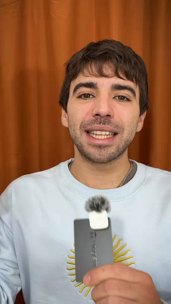 The image features a man wearing a blue sweater, holding up an electronic device with a camera attached to it. The background of the photo has orange curtains, which adds a warm and inviting atmosphere to the scene. The caption provided after the image is in Spanish, discussing the various uses for the device being held by the man.