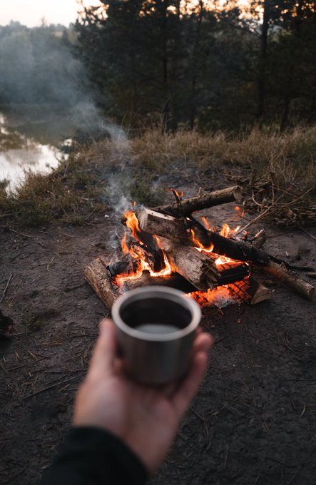First-person perspective photo of a hand holding a metal cup of coffee in front of a crackling campfire by a lakeside at dusk.