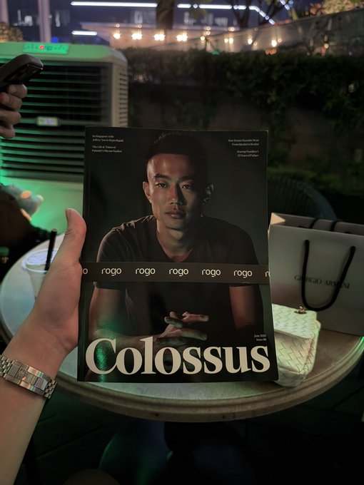 A hand holds a 'ROGO' magazine titled 'Colossus' featuring a young Asian man, with a Giorgio Armani shopping bag visible on the table behind it.