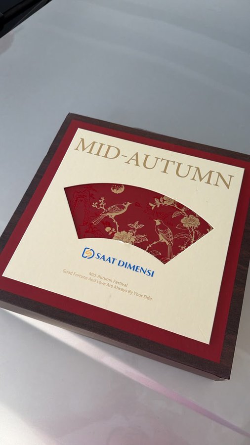 A Mid-Autumn Festival gift box from 'SAAT DIMENSI' featuring a red fan-shaped design with gold floral and bird motifs, with text reading 'Mid-Autumn Festival, Good Fortune And Love Are Always By Your Side'.