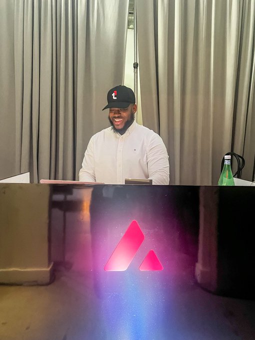 A smiling bearded man wearing a black baseball cap with a red logo and a white shirt sits at a sleek black desk illuminated by a glowing pink and blue triangular logo, with a green water bottle beside him.