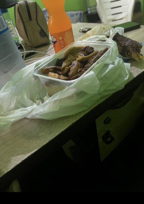 A takeout meal on a wooden desk featuring a container of roasted vegetables and meat, an orange Fanta bottle, a water bottle, and a side dish, all resting on a white plastic bag.