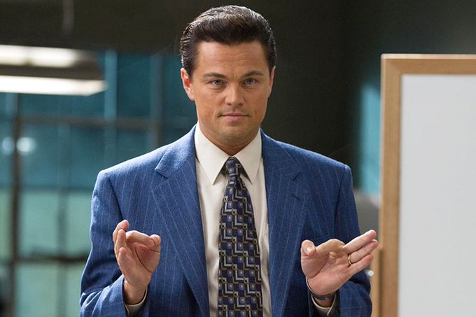 A man in a blue pinstripe suit, white shirt, and patterned tie gestures with his hands while speaking in what appears to be an office setting.