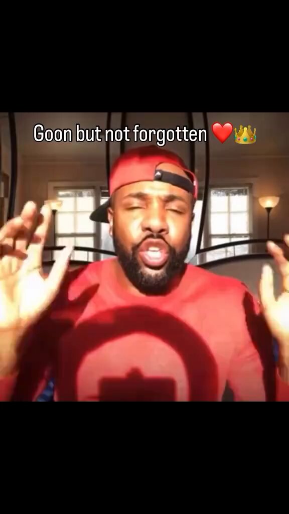 The image features a man wearing a red shirt, with his hands raised as if he's making an argument or expressing frustration. He has a black hat on backwards, adding a touch of humor to the scene. The background includes a window that allows natural light into the room and a lamp, creating a cozy atmosphere. The caption below the image reads "Gm $niggabutt nation", which is likely a reference to the popular meme or trend involving the phrase "Gm but not forgotten".