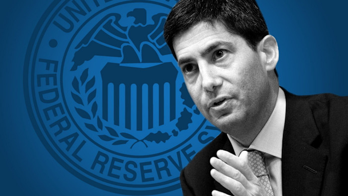 A black-and-white photo of a man in a suit speaking, overlaid on a blue background featuring the seal of the Federal Reserve.