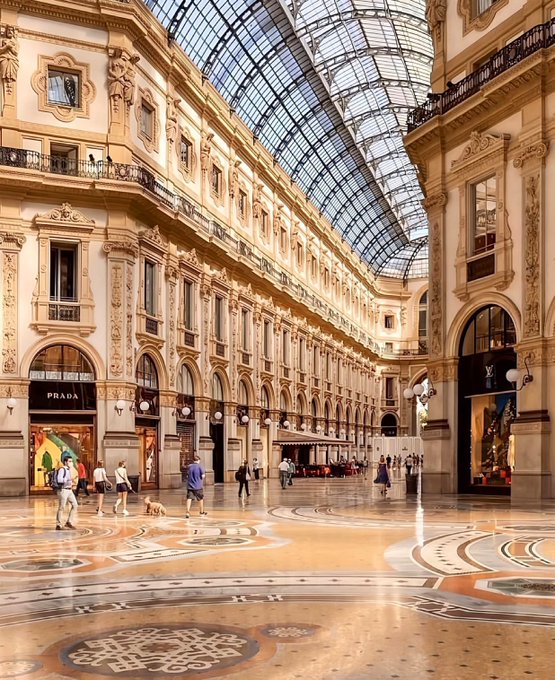 An elegant interior view of the Galleria Vittorio Emanuele II in Milan, featuring ornate architecture, a vaulted glass ceiling, a Prada storefront, and visitors walking on the intricate mosaic floor.