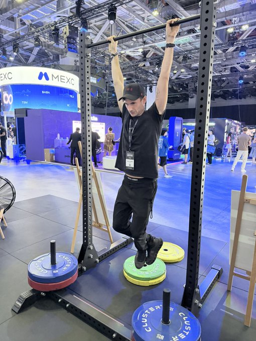 A man performs a pull-up on a metal rig at what appears to be a fitness convention, with a MEXC booth visible in the background and 'CLUSTER' branded weight plates on the floor.