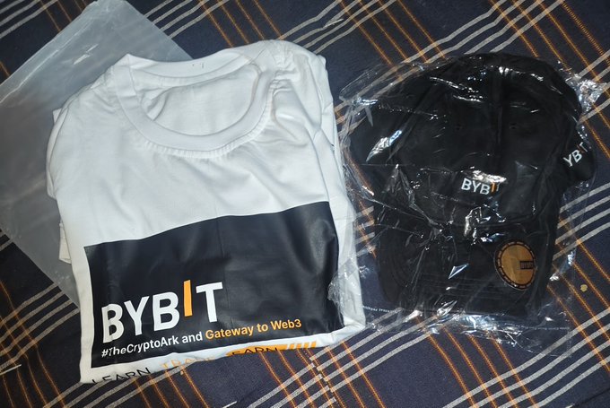 The image features a white t-shirt and a black baseball cap sitting next to each other on a bedspread, both wrapped in plastic bags. The t-shirt has a "Bybit" logo on it, indicating that it might be a gift from Bybit. The post about receiving the gift by @Bybit_Official emphasizes gratitude towards the sender of the gift and mentions the hashtag #JihadWeb3 to highlight the connection between the gift and the hashtag.
