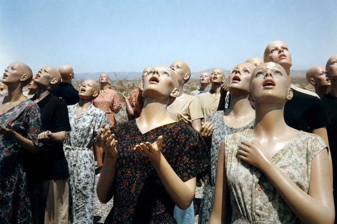 A surreal outdoor scene showing a crowd of bald, mannequin-like figures in floral dresses looking upward with open mouths, set in a desert landscape under a clear sky.