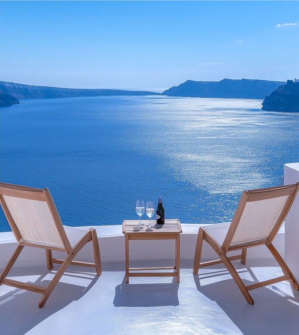 Two wooden lounge chairs and a small table with a bottle of wine and two glasses sit on a white terrace overlooking a stunning blue sea and cliffs, characteristic of Santorini.