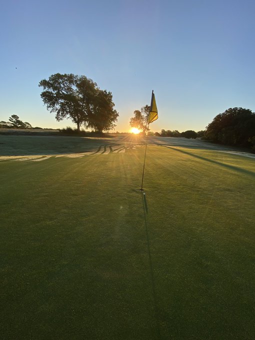 A scenic golf course at sunrise or sunset with a yellow flag marking the hole, a tree silhouetted in the background, and the sun creating a bright glow on the horizon.