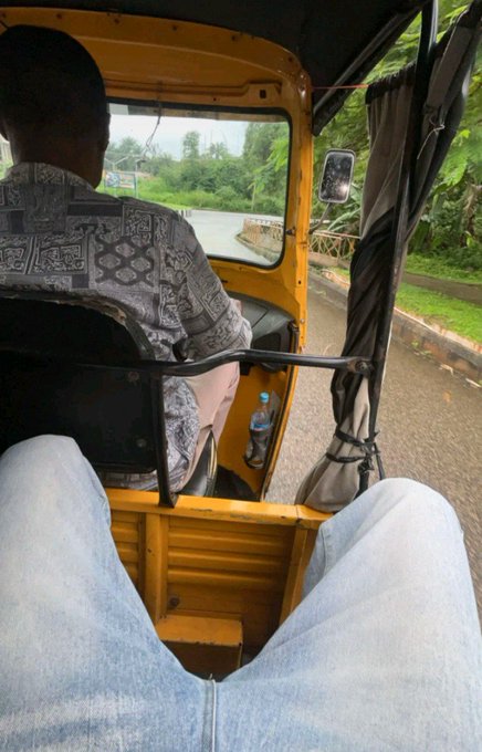 A first-person view from inside a tuk-tuk shows a driver in a patterned shirt navigating a wet, paved road lined with greenery.