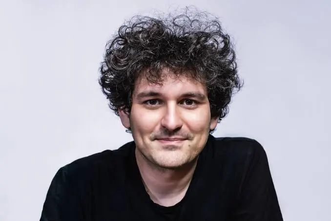 The image features a man with curly hair wearing a black shirt, looking directly at the camera with a smile. The background appears to be white or light gray, which contrasts with the man's dark attire. There are no other objects or people visible in the image.