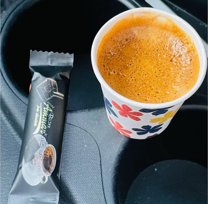 A paper cup filled with espresso sits in a car cup holder next to a stick-pack of 'Alio American Coffee' with 'MILK' flavor.