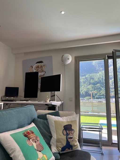 A bright living room showcases two Bored Ape NFT-themed decorative pillows—one featuring a crowned ape and the other a top-hatted ape—echoing the "Bored Ape is such a vibe" caption, complemented by a matching ape artwork hanging above a dual-monitor desk setup overlooking a scenic balcony.