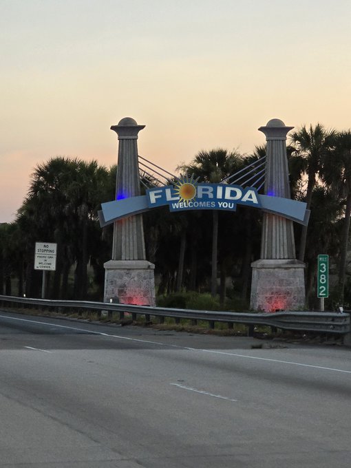 A 'FLORIDA WELCOMES YOU' sign stands at a roadside border at dusk, flanked by tall white pillars and palm trees, with mile marker '302' visible on the right.