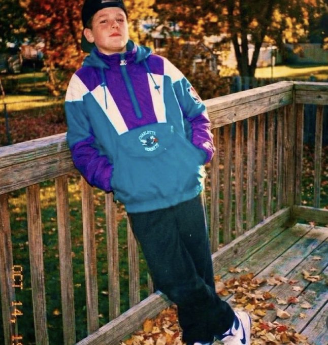 A vintage photograph of a young boy wearing a blue, purple, and white Charlotte Hornets Starter jacket and a backwards cap, posing on a wooden deck surrounded by autumn leaves.