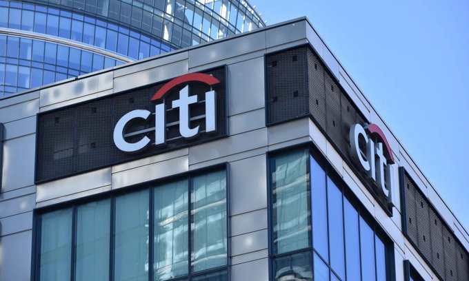 The exterior corner of a modern glass office building displays a large 'citi' logo with the signature red arc on a metallic panel against a clear blue sky.