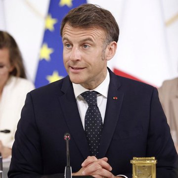 French President Emmanuel Macron sits at a meeting table with EU and French flags behind him. The image accompanies a claim that France’s parliament advanced a 31.4% capital-gains tax on Bitcoin and crypto-to-stablecoin swaps, though no tax figures or related text are visible in the photo itself.