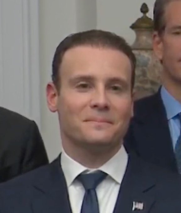A close-up photo of Emmanuel Macron, President of France, wearing a dark suit and tie with a small French flag pin on his lapel, with another man partially visible behind him.