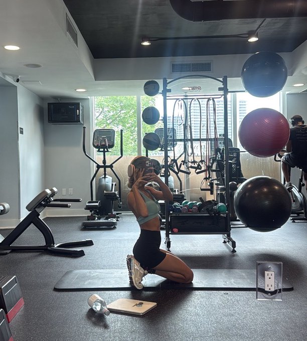 A woman in athletic wear kneels on a mat in a gym, holding up her phone, surrounded by workout equipment including exercise balls and a rack of medicine balls.