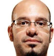 A close-up photo of a bald man with glasses, a goatee, and sideburns looking directly at the camera.