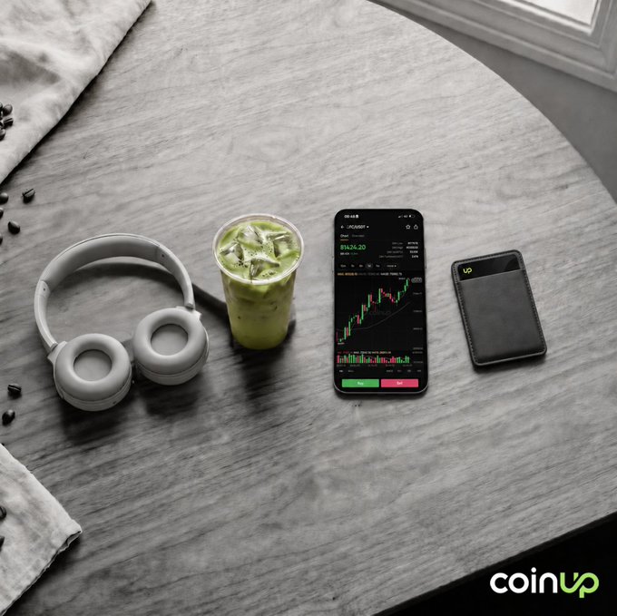 The image shows a wooden table with various items placed on top of it, including headphones, a cell phone displaying stock market data, a cup of green tea, and a wallet. The text "coinup fam" appears at the bottom left corner of the image.