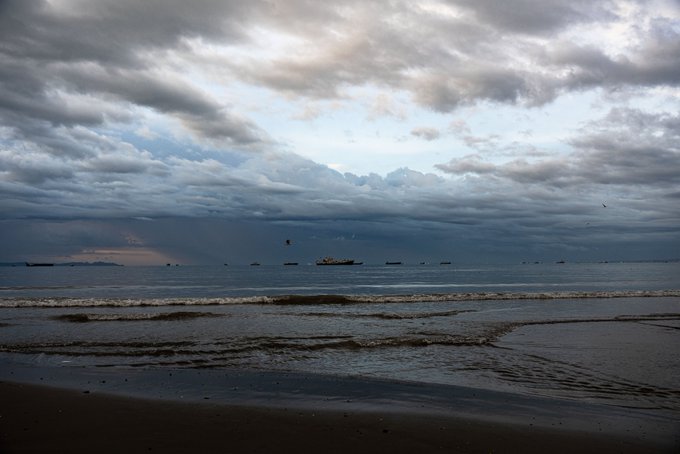 Moody coastal scene of a beach at dusk or dawn, with overcast clouds, small boats on the horizon, and a faint sun visible near the water.