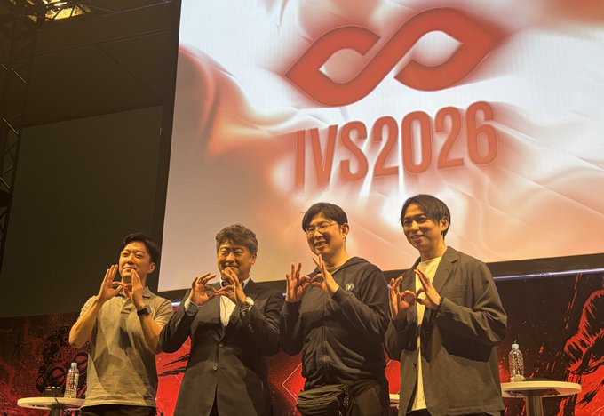 Four men pose on stage in front of a large screen displaying the "IVS2026" logo, each forming an "OK" hand gesture. The setting corresponds to the IVS2026 session mentioned in the post, featuring Professor Kihara, Mr. Saito, and Professor Matsukura, with the speaker set to deliver two presentations the following day.
