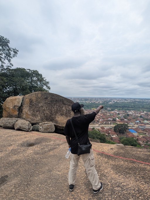 A person wearing a black shirt, cap, and crossbody bag stands on a rocky outcrop, pointing out over a sprawling cityscape under an overcast sky.