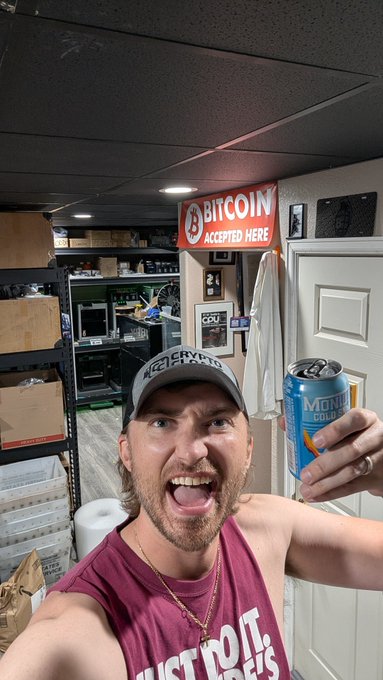 A shirtless man wearing a "Just Do It" Nike cap takes an excited selfie while holding up a tall can of "Michelob Ultra" beer, celebrating his appreciation for whoever treated him to a drink, with a "Bitcoin Accepted Here" sign visible in the background of the shop.