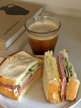 A sandwich cut in half with ham, cheese, and cucumber sits on a white plate next to a glass of tea, with a book titled 'Les Misérables' in the background.