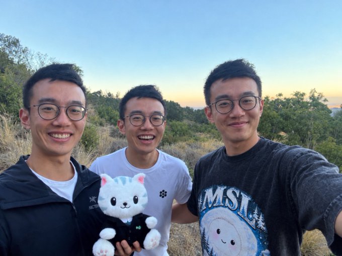 A selfie of three smiling young Asian men wearing glasses, standing outdoors at sunset, with one holding a white cat plush toy.