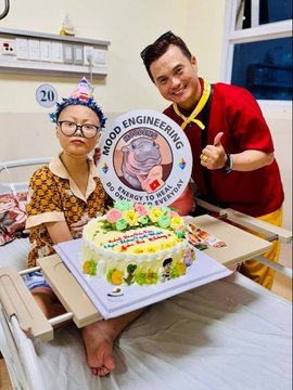 A man in a red shirt presents a 'Mood Engineering' themed birthday cake to an elderly woman wearing glasses who is sitting in a hospital bed with party decorations.