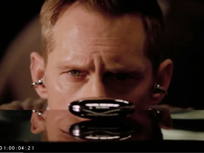 A close-up still of a man with short blonde hair wearing earbuds, looking down at a reflective black object, with a timecode '01:00:04:21' visible at the bottom left.