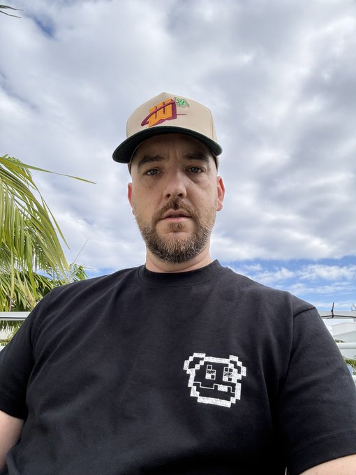 A selfie of a man wearing a black t-shirt with a pixelated graphic and a baseball cap with a Mountain Dew logo, standing outdoors with palm trees and cloudy sky in the background.