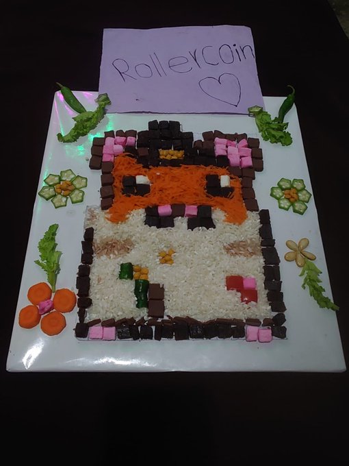 A pixel-art style sushi cake depicting a cat character, bordered by chocolate pieces and garnished with vegetables, with a purple note reading "RollerCoin" and a heart symbol.