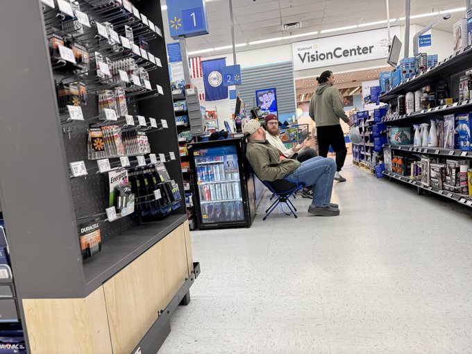 A Walmart store interior showing two people sitting in camping chairs in the aisle near the 'VisionCenter' section, with 'Walmart+' signage and product shelves visible.