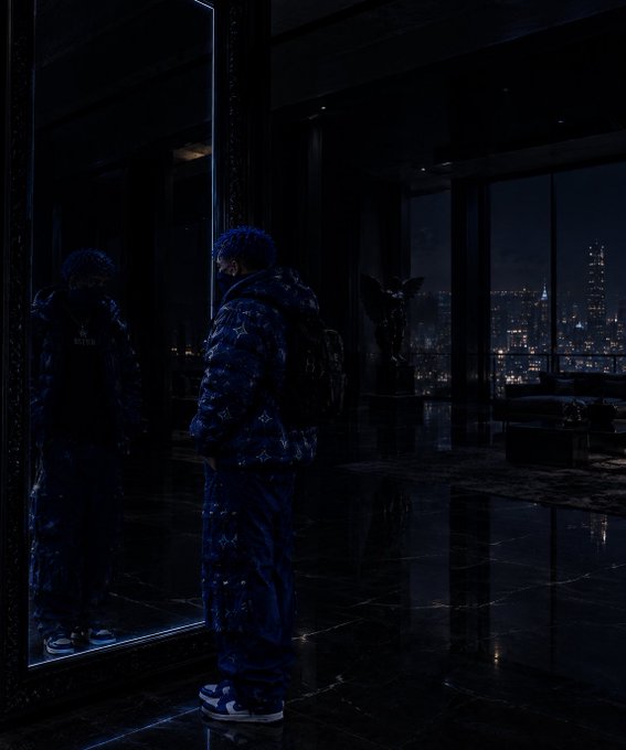 A figure wearing a blue Louis Vuitton monogram puffer jacket and matching pants stands in front of a large mirror in a dimly lit luxury room overlooking a city skyline at night.