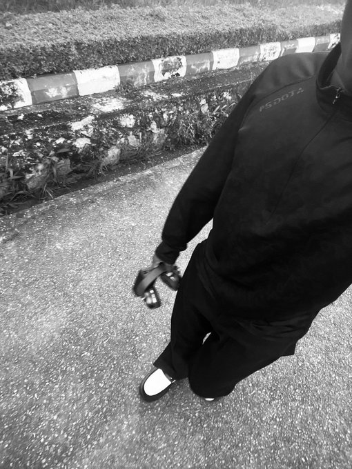 A black and white photo of a person wearing a black tracksuit with 'PSGOTA' written on the chest, carrying a small object in their hand while walking on a paved surface next to a curb.