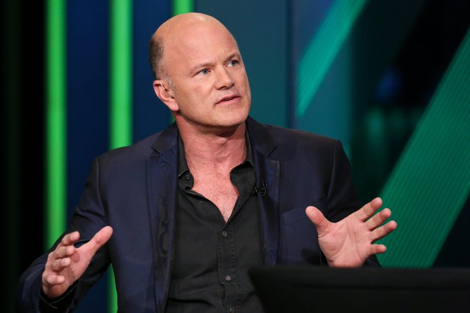 A bald man in a dark blue blazer and black shirt gestures expressively with both hands open while speaking on a stage with green vertical lighting in the background.