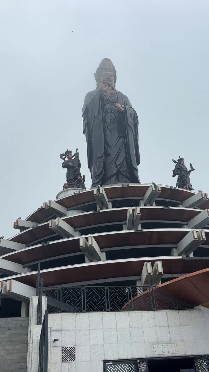 The image features a large statue of a man, possibly a Buddhist monk or a deity, situated atop a building. There are three smaller statues placed around the main figure, adding to its grandeur. The scene appears to be set against an overcast sky, which adds a sense of solemnity and tranquility to the overall atmosphere.