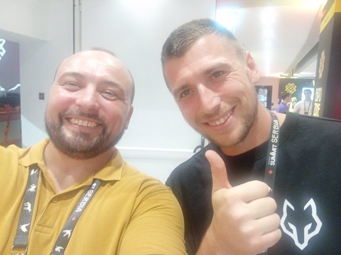 Two men pose for a selfie at what appears to be a gaming convention, both wearing lanyards labeled 'SUMMIT SERBIA'.