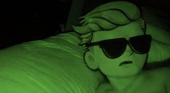 A person with short blonde hair wearing dark sunglasses lies on a white pillow in a dimly lit room bathed in green light.