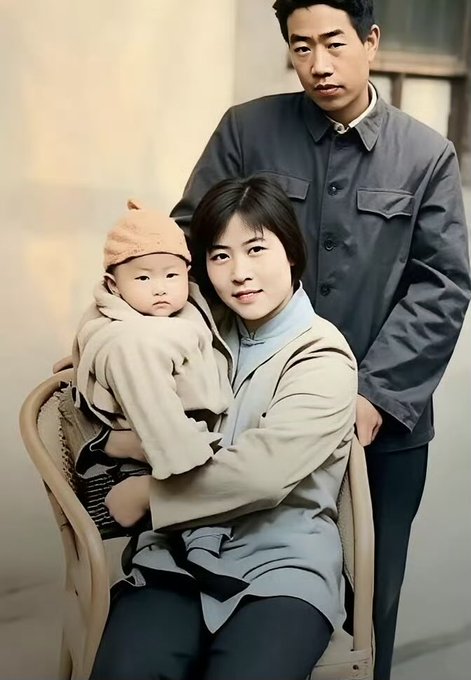 A vintage-style family portrait showing a man in a gray Mao suit standing behind a seated woman holding a baby wearing a beige knit hat.